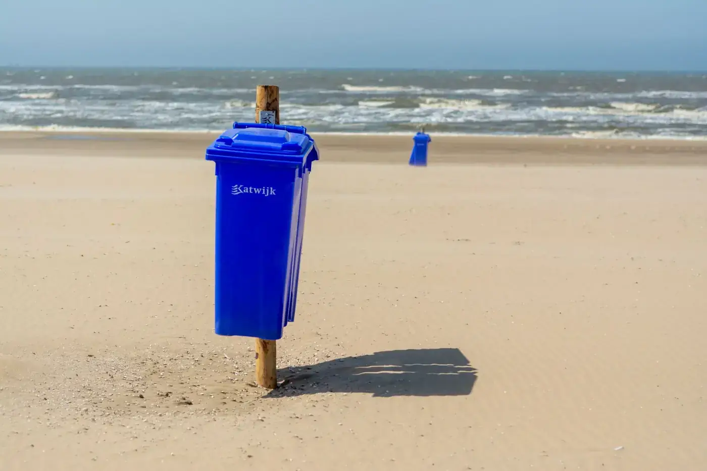 Sunlit beach with blue garbage bins by the sea in Katwijk aan Zee, Netherlands.