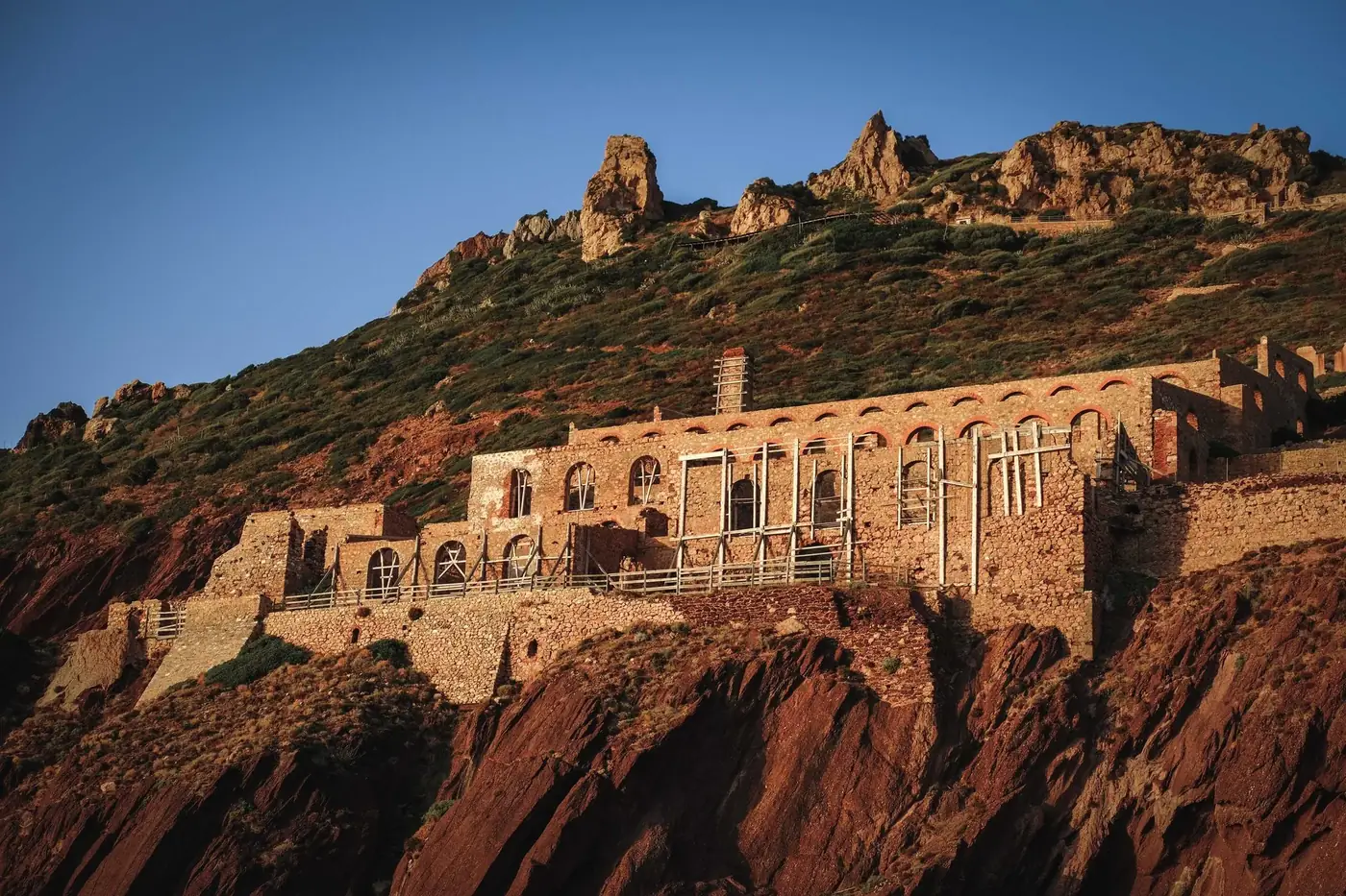 Exterior of ancient abandoned building of Laveria Lamarmora mine built of rocky cliff slope against cloudless blue sky i