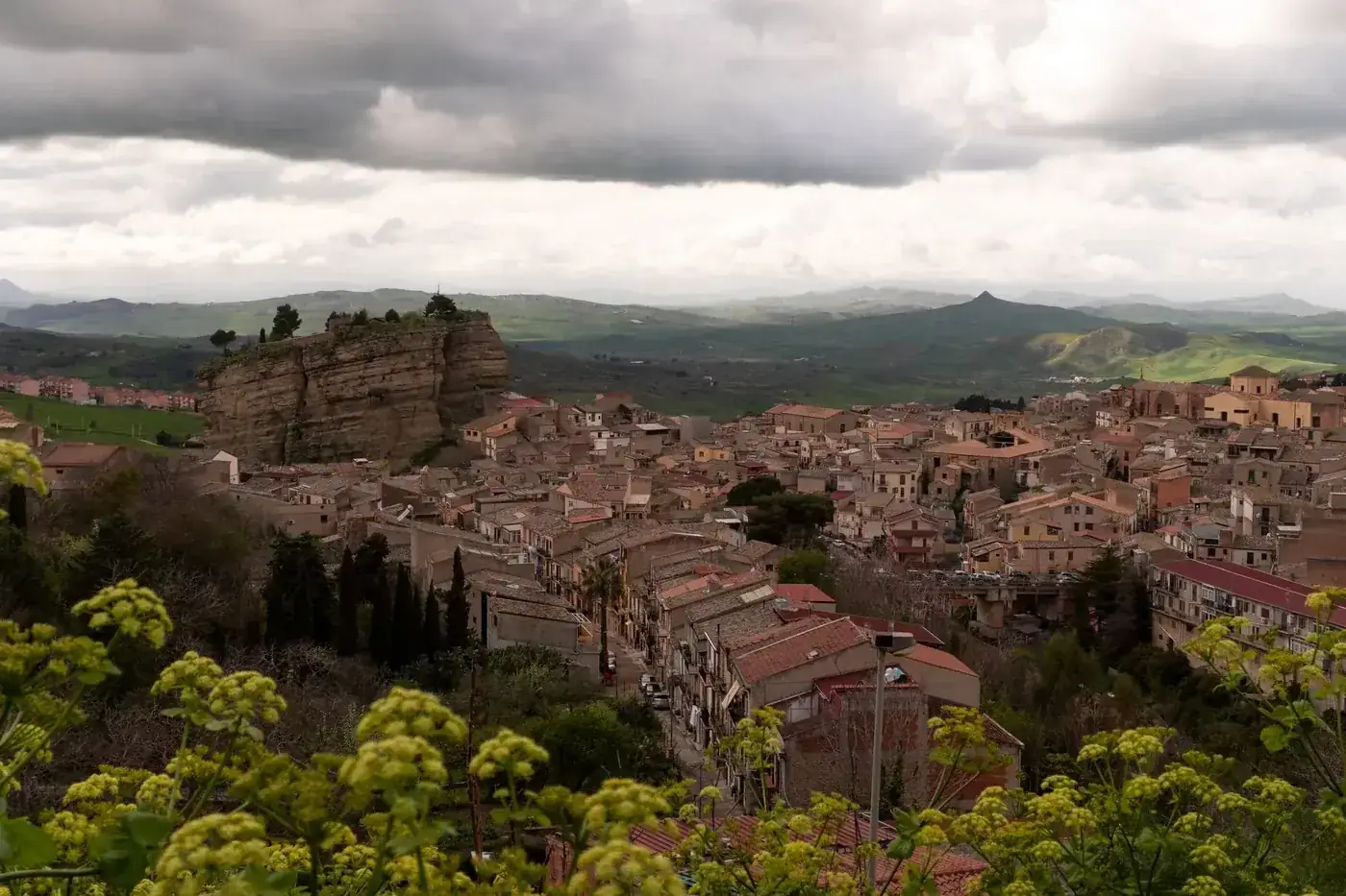 Aerial view of a quaint Sicilian town, with rolling hills and cloudy skies in the background.