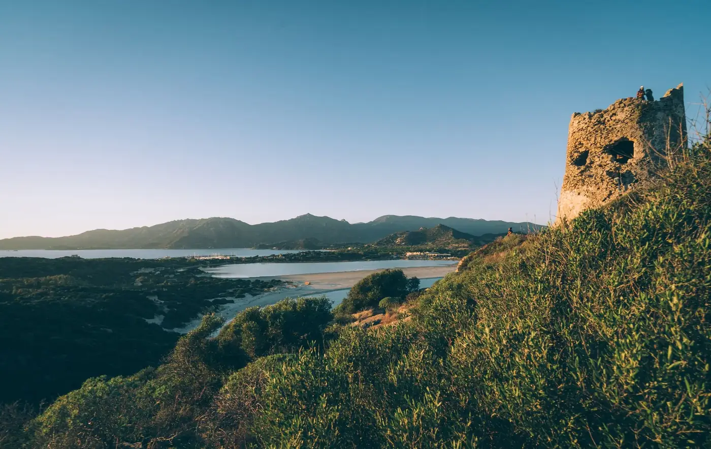 Stunning landscape of a coastal bay with mountains and an ancient tower at sunset.