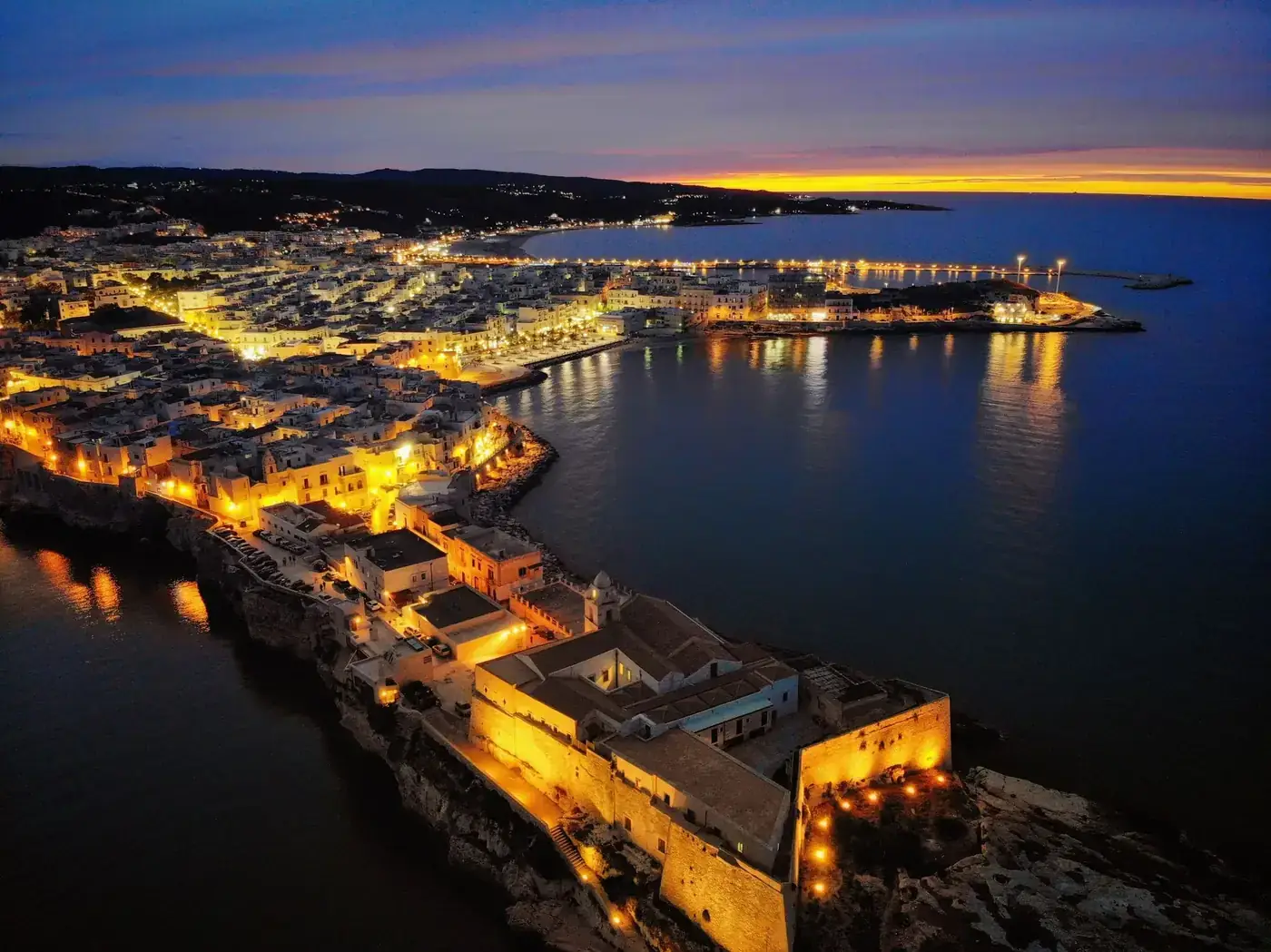 Stunning aerial shot of illuminated Vieste, Italy, with coastal view during sunset.