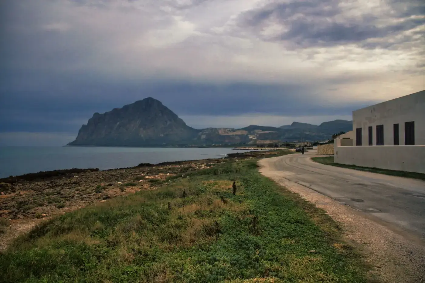 Tranquil coastal road with Monte Cofano in Sicily, Italy under cloudy skies.