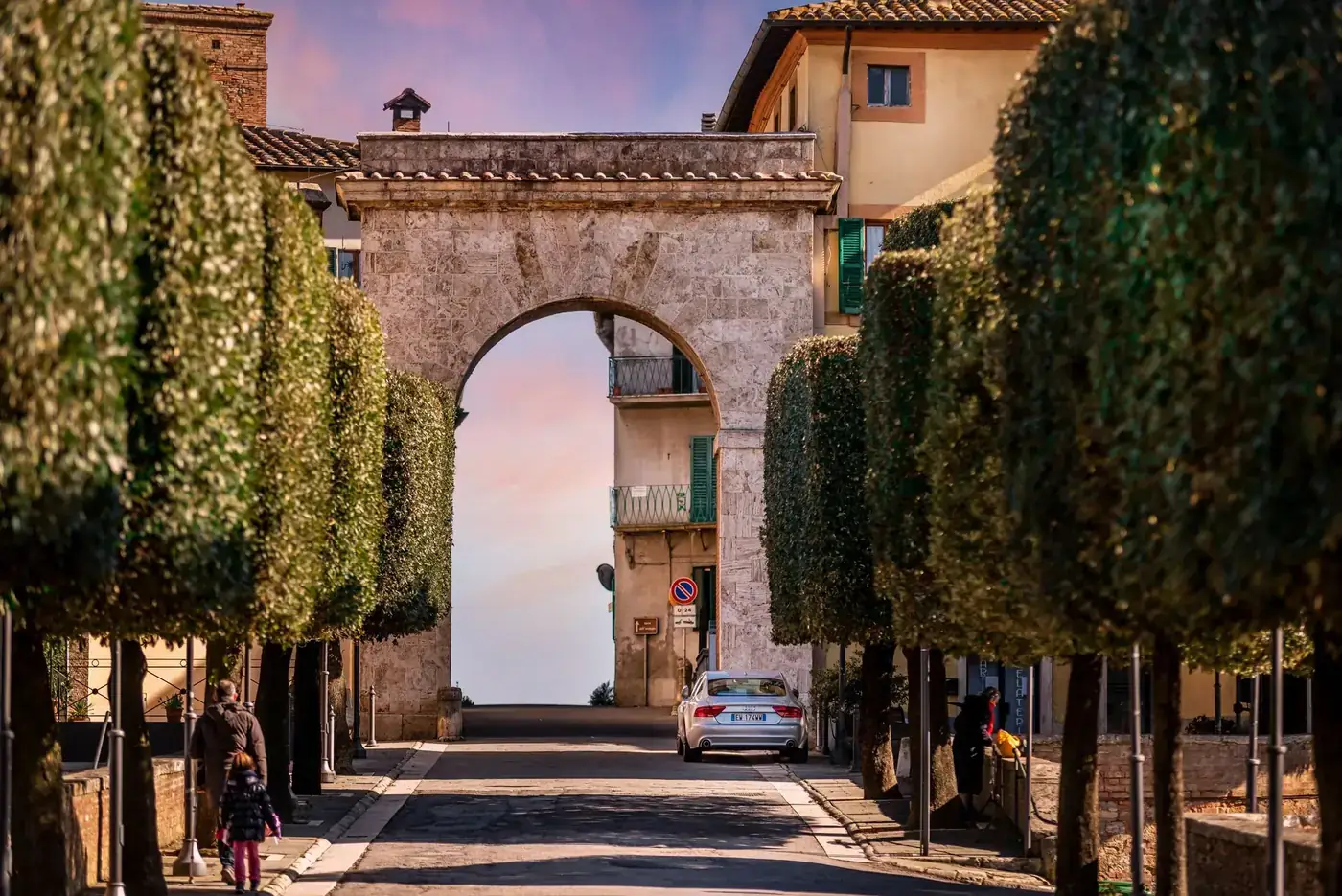 Narrow cobblestone street amidst lush green trees leading to stone arch against sunset sky in old town of Chianciano Ter