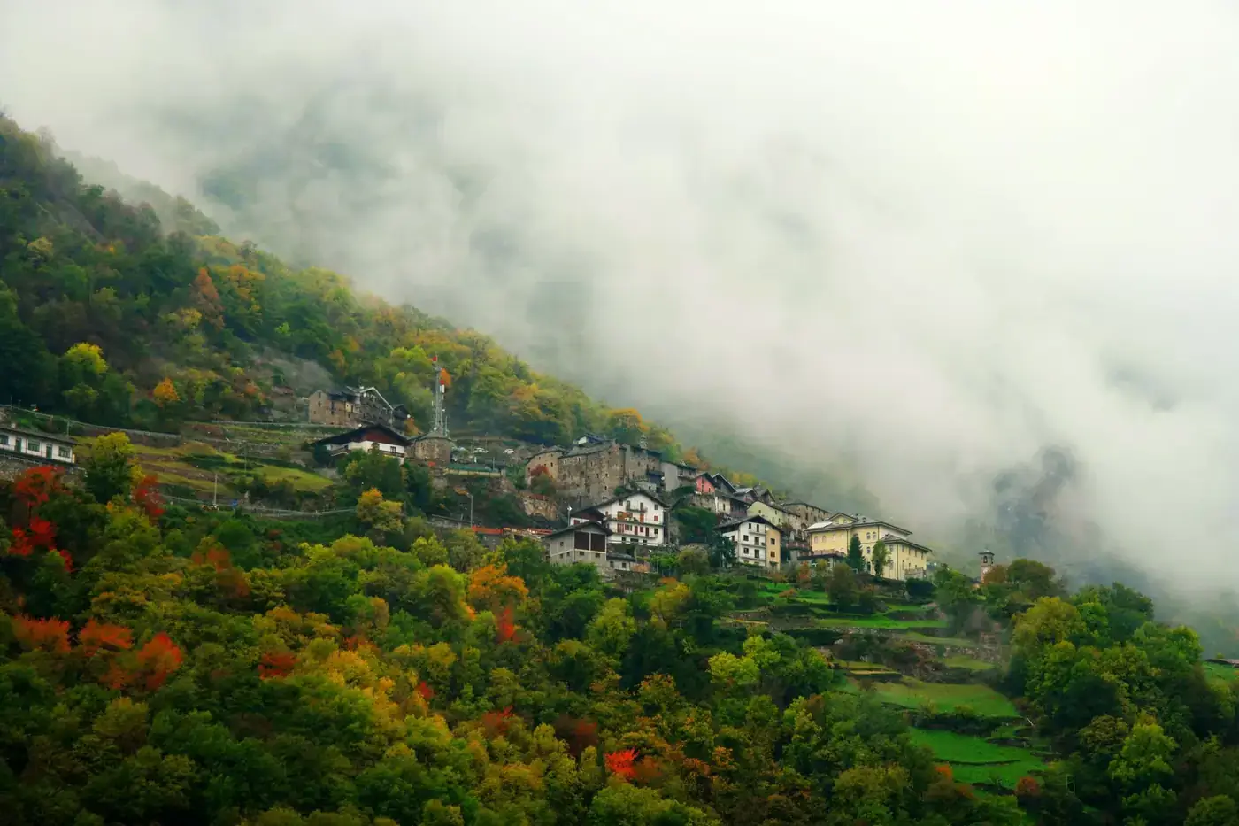 Foggy autumn scene of a hillside village in Issime, Italy, with vibrant fall colors and mist.