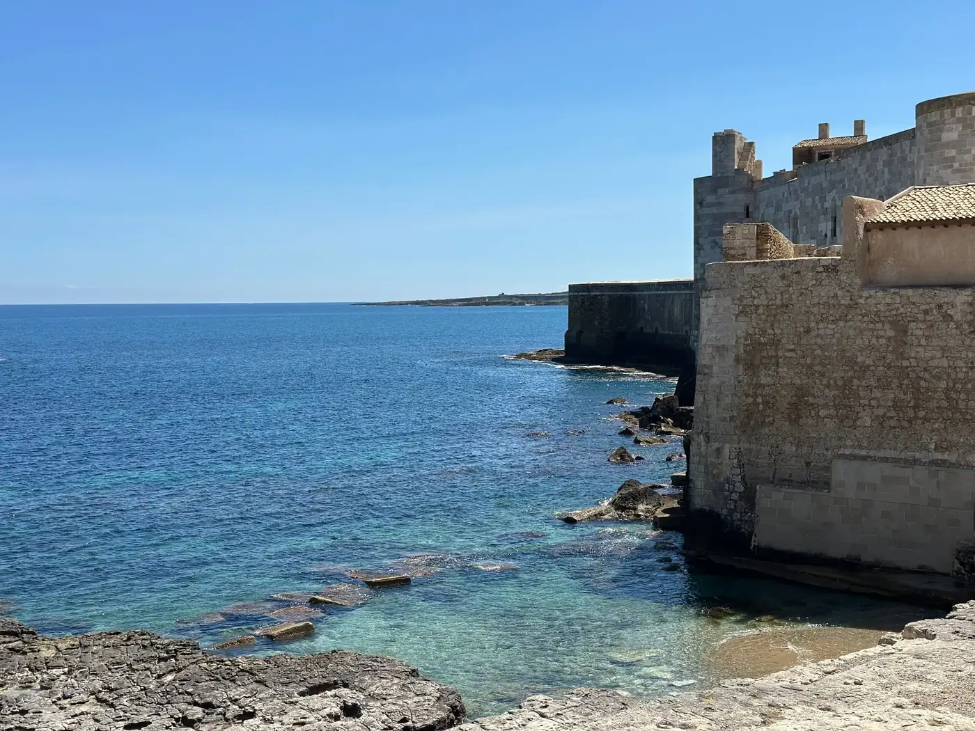 Scenic view of Ortigia's coastline in Sicily with historical stone buildings against clear blue waters.