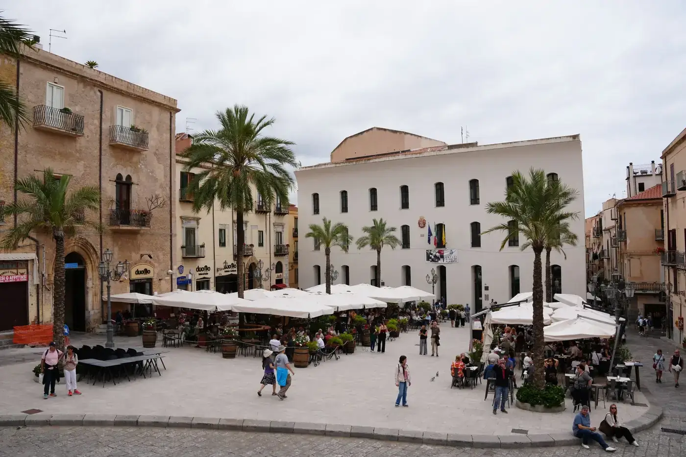 Busy Piazza Del Duomo in Cefalù, Italy, bustling with people and cafes under a cloudy sky.