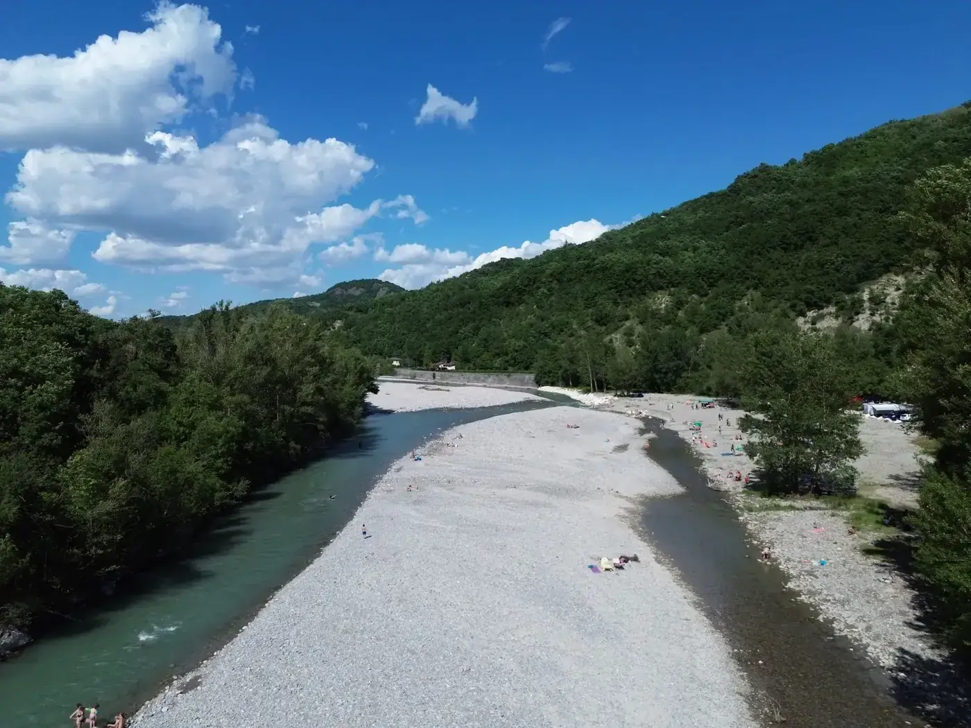 Aerial view of a river, pebble beach, and lush greenery under a bright summer sky in Italy.