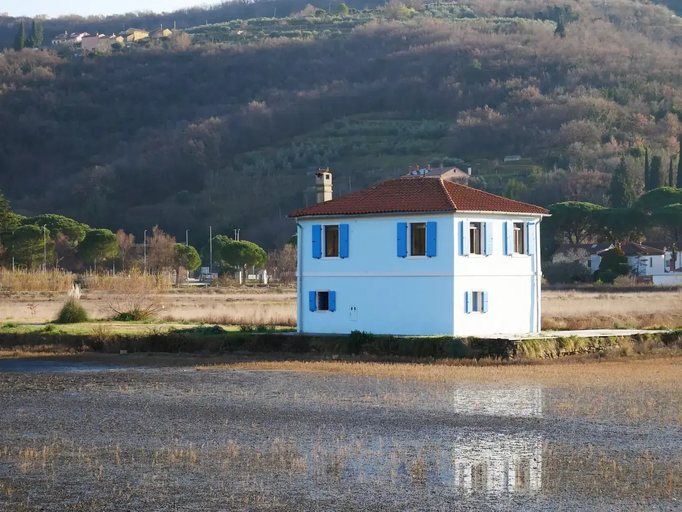 A serene landscape featuring a blue house surrounded by fields and hills under a blue sky.