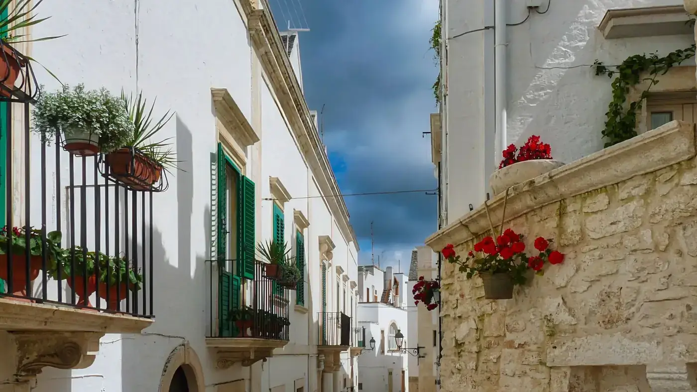 Vibrant flowers adorn balconies in the whitewashed streets of Ostuni, Italy, under a dramatic sky.