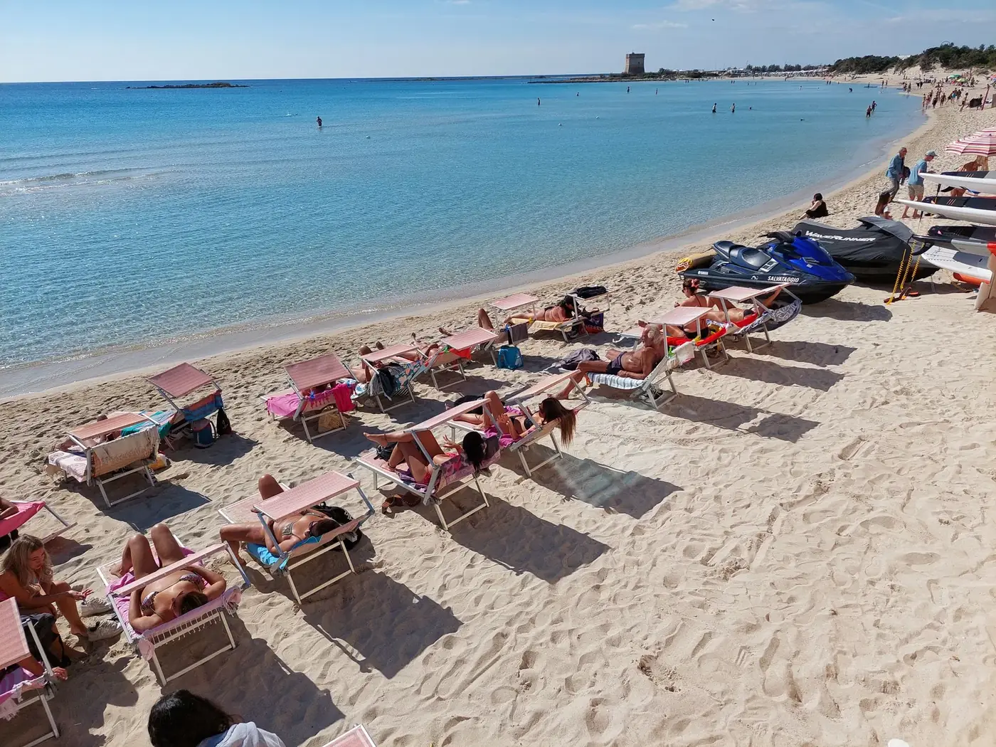 A vibrant summer day at a sandy beach with sunbathers lounging and jet skis nearby.
