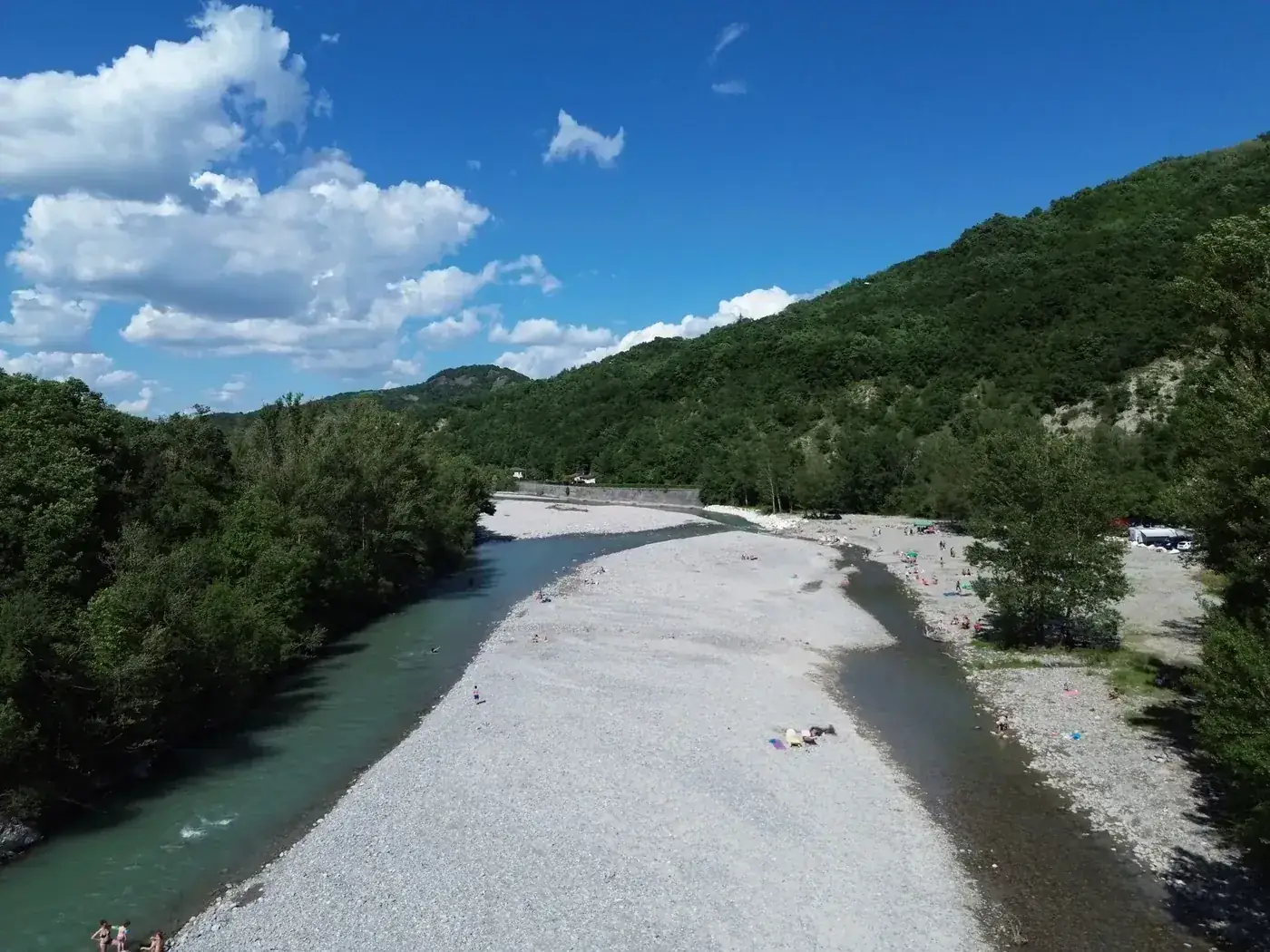 Aerial view of a river, pebble beach, and lush greenery under a bright summer sky in Italy.