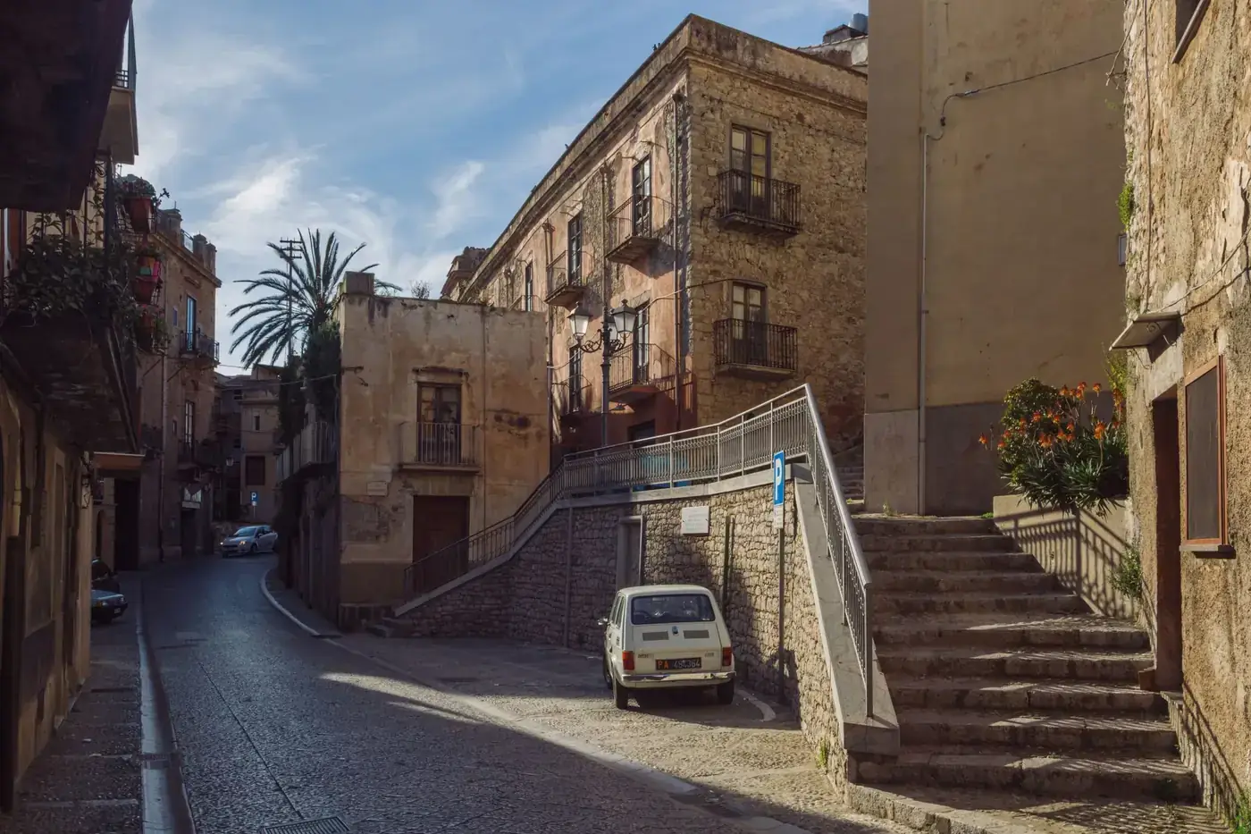 Sunlit street view in Caccamo, Sicily with classic architecture and cars.